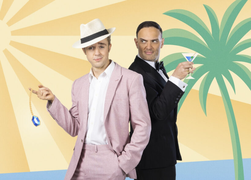 One man in a pink suit and fedora holding a diamond on a chain standing beside another man in a black tie suit holding a martini