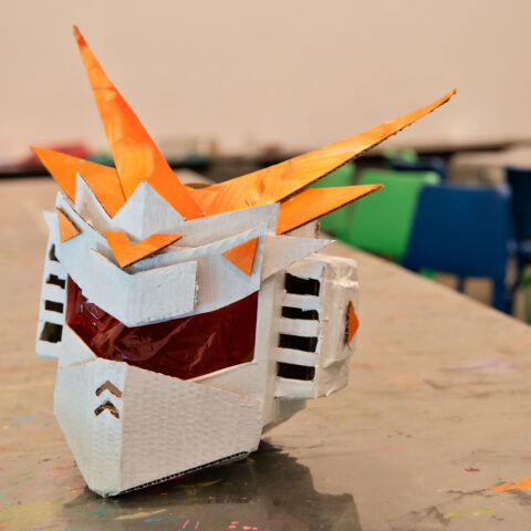 A robot helmet made from painted cardboard and cellophane sits on an art making table 