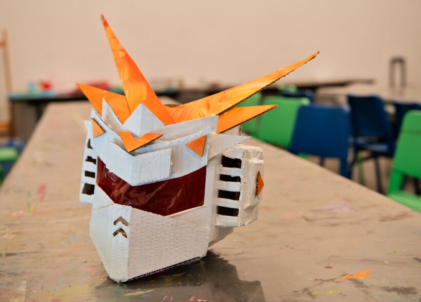 A robot helmet made from painted cardboard and cellophane sits on an art making table 
