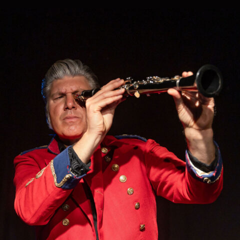 A man in a red office uniform looks through a clarinet like a telescope