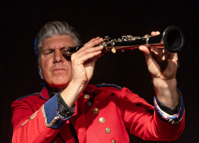 A man in a red office uniform looks through a clarinet like a telescope
