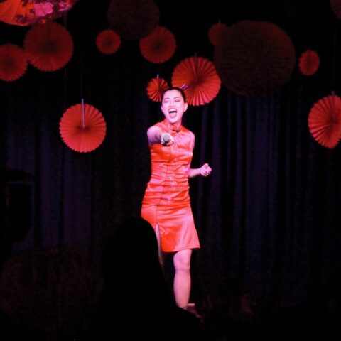 A woman in a red dress passionately sings on stage, holding a microphone. Red paper decorations hang in the background. The atmosphere is vibrant and energetic.