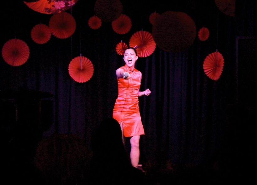 A woman in a red dress passionately sings on stage, holding a microphone. Red paper decorations hang in the background. The atmosphere is vibrant and energetic.