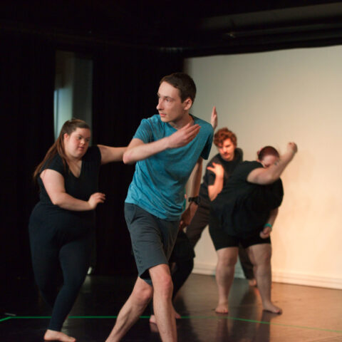 A young man dancing on stage in a blue t-shirt while others dance behind him. 