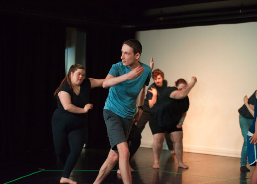 A young man dancing on stage in a blue t-shirt while others dance behind him. 