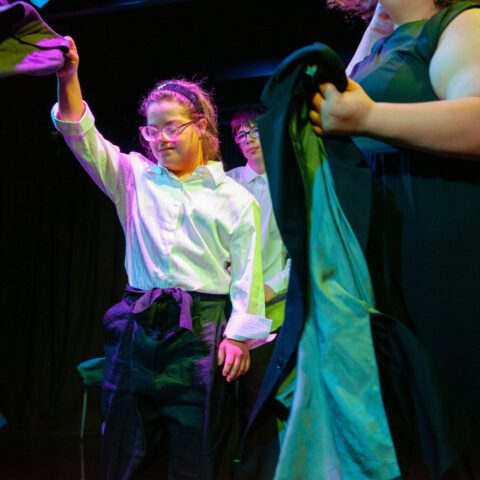 Young people perform during a Restless Dance Theatre end-of-year showing. A young person in a white button up top dances while throwing a jacket rhythmically in the air. In the foreground other young people move with their jackets on stage.