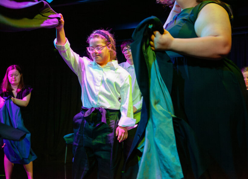 Young people perform during a Restless Dance Theatre end-of-year showing. A young person in a white button up top dances while throwing a jacket rhythmically in the air. In the foreground other young people move with their jackets on stage.
