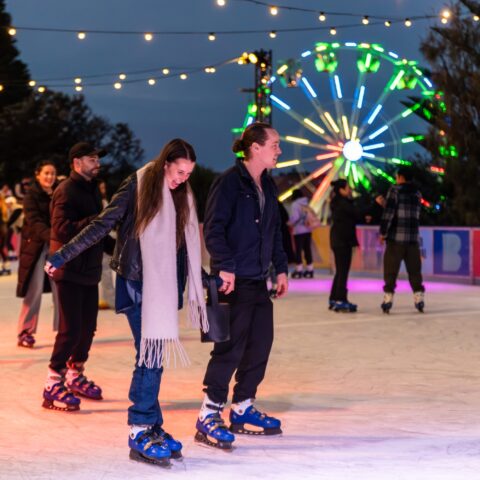 People enjoy ice skating at night under colorful lights and string lanterns, with a neon-lit Ferris wheel in the background, creating a festive atmosphere.