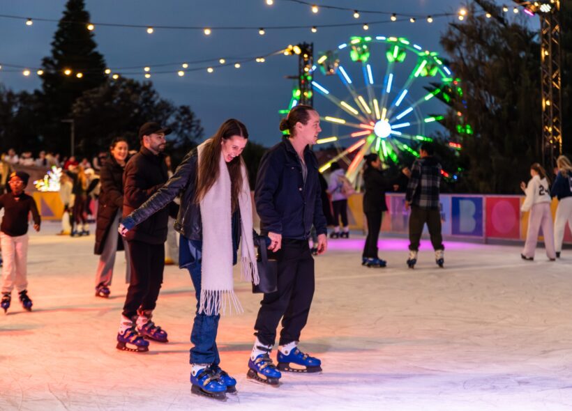 People enjoy ice skating at night under colorful lights and string lanterns, with a neon-lit Ferris wheel in the background, creating a festive atmosphere.