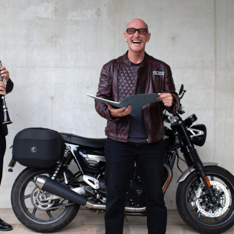 Two men stand next to a motorcycle, one in a tuxedo with a clarinet and another in a leather jacket holding an open book