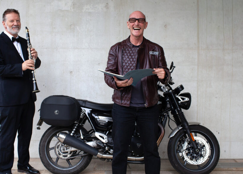 Two men stand next to a motorcycle, one in a tuxedo with a clarinet and another in a leather jacket holding an open book