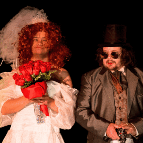 A person in a white wedding dress holds red roses beside another in a dark suit and top hat. Both exude a theatrical vibe against a black backdrop.