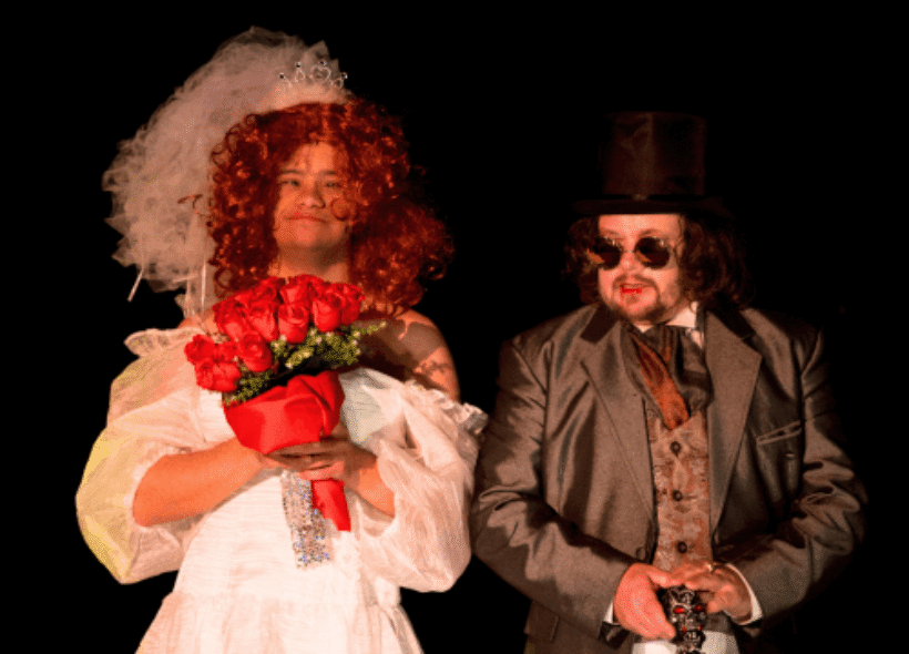 A person in a white wedding dress holds red roses beside another in a dark suit and top hat. Both exude a theatrical vibe against a black backdrop.