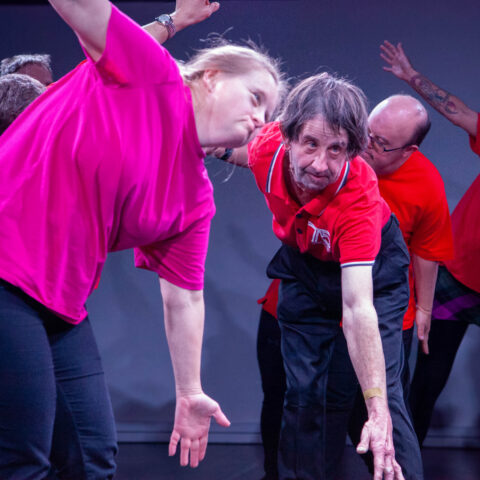 An older man in a red shirt and women in a pink shirt mimic each other's dance move as other dancers move behind them. 