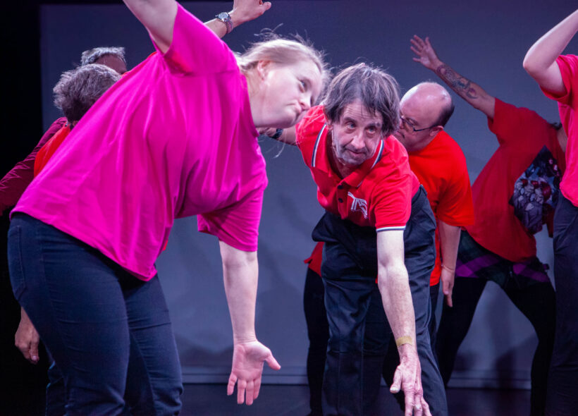 An older man in a red shirt and women in a pink shirt mimic each other's dance move as other dancers move behind them. 