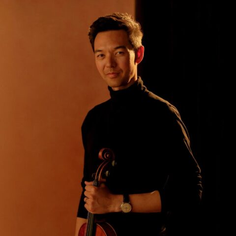 A person stands holding a violin against a warm, brown backdrop. They wear a black shirt, and their expression is calm and confident.