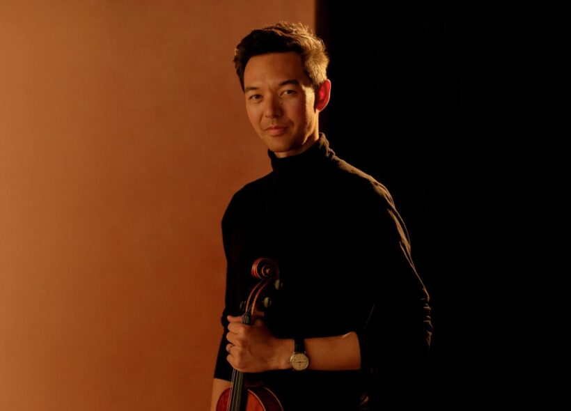 A person stands holding a violin against a warm, brown backdrop. They wear a black shirt, and their expression is calm and confident.