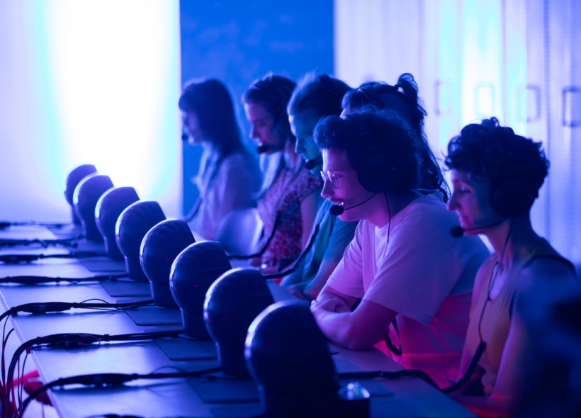 A row of people wearing headsets sit at computers in a dimly lit room with blue and pink lighting. 