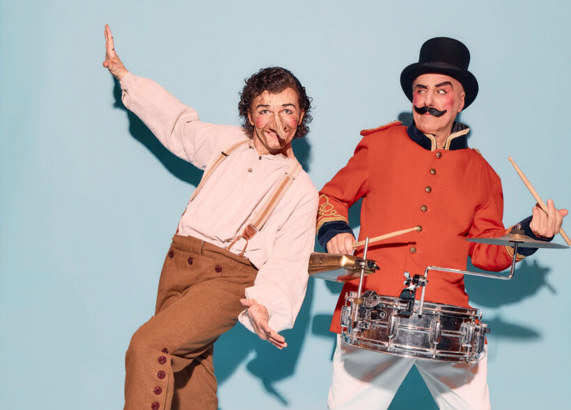 Man made out of wood and pinocchio's nose next to a red coat man in a toop hat and big moustache 