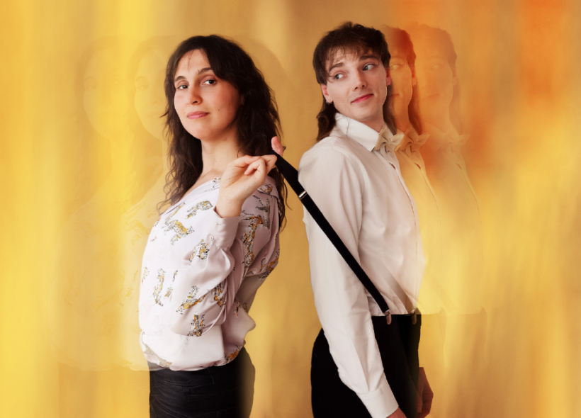 Two people stand back-to-back against a warm, blurred background. One wears a patterned shirt. The other is in a bow tie and suspenders, looking to the side. 