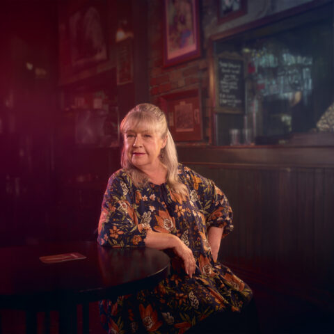A woman with long, light hair and a floral dress sits at a dimly lit bar, exuding a contemplative mood. Warm lighting and framed pictures create a cozy, nostalgic atmosphere.