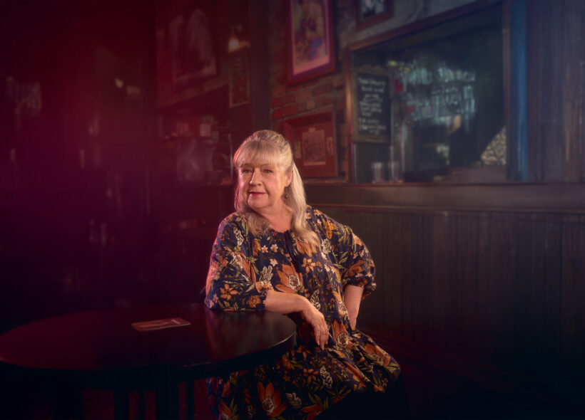 A woman with long, light hair and a floral dress sits at a dimly lit bar, exuding a contemplative mood. Warm lighting and framed pictures create a cozy, nostalgic atmosphere.