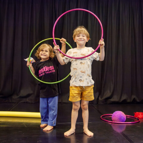 Two children participating in a Restless Dance Theatre WONDER class smile while holding hula hoops in a dance studio. Colourful movement props are arranged around them on the floor.