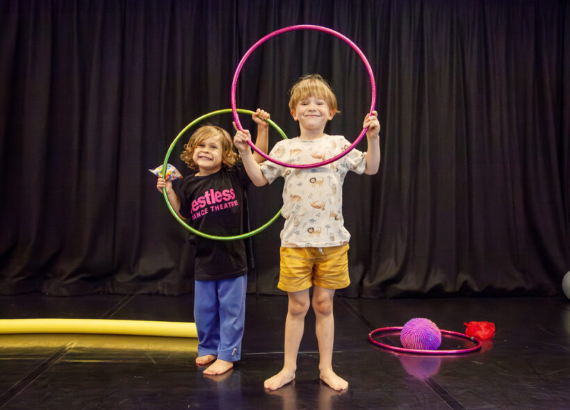 Two children participating in a Restless Dance Theatre WONDER class smile while holding hula hoops in a dance studio. Colourful movement props are arranged around them on the floor.