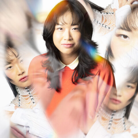 A woman in an orange sweater is centered, surrounded by multiple blurred and overlapping images of herself against a white background.
