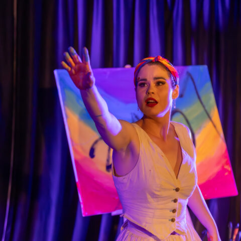 A performer in a white dress reaches toward the audience on a dimly lit stage, standing in front of a brightly coloured painted canvas under purple theatrical lighting.