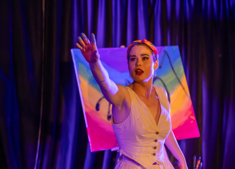 A performer in a white dress reaches toward the audience on a dimly lit stage, standing in front of a brightly coloured painted canvas under purple theatrical lighting.