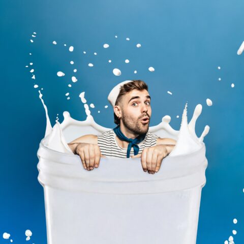 A man in a striped shirt appears to be sitting in a container of milk, with milk splashing around him, against a blue background. 