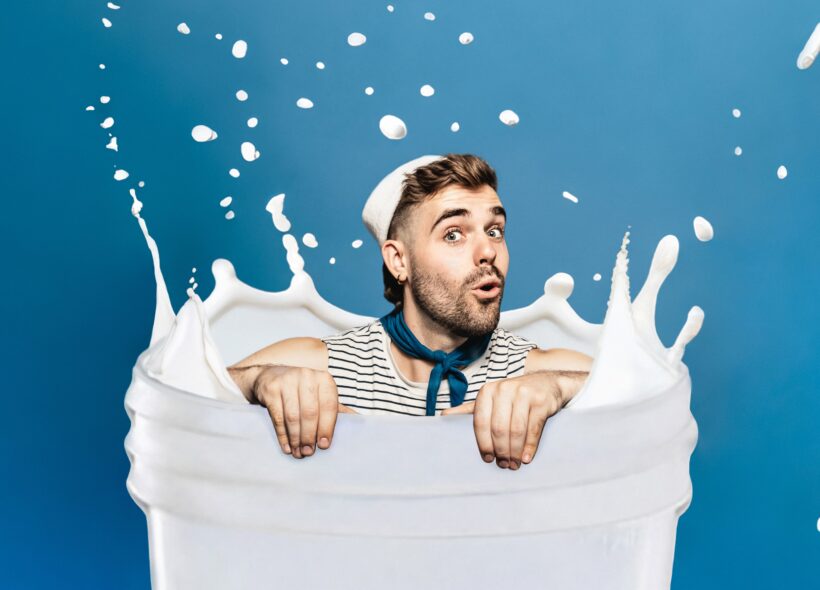 A man in a striped shirt appears to be sitting in a container of milk, with milk splashing around him, against a blue background. 