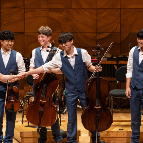 A group of young musicians joyfully playing violins and a cello together in a bright, lively setting