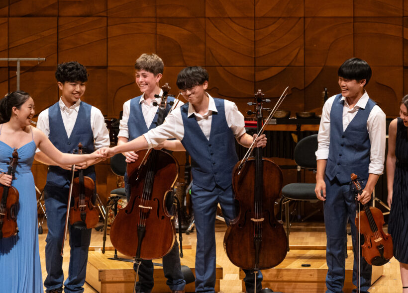 A group of young musicians joyfully playing violins and a cello together in a bright, lively setting