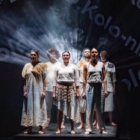 A group of people standing proudly side by side on a brightly lit stage, with words projected across them