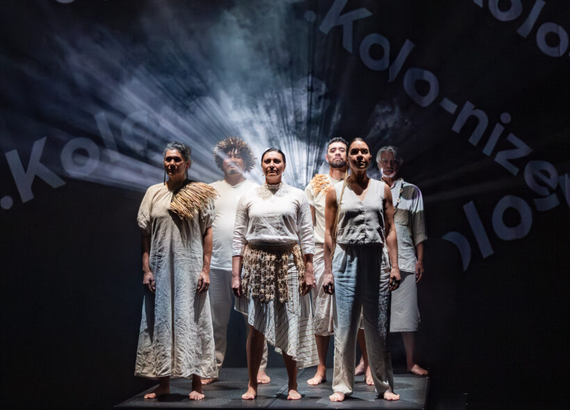A group of people standing proudly side by side on a brightly lit stage, with words projected across them