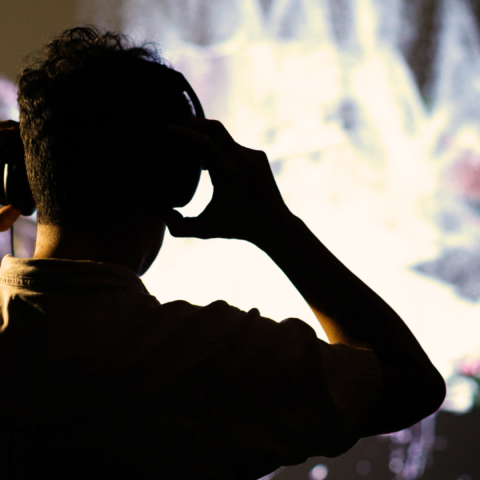A photo of a person from behind. The person is adjusting headphones over their ears and watching a video artwork. 