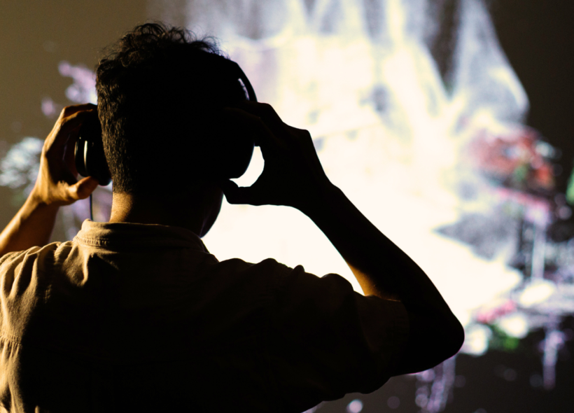 A photo of a person from behind. The person is adjusting headphones over their ears and watching a video artwork. 