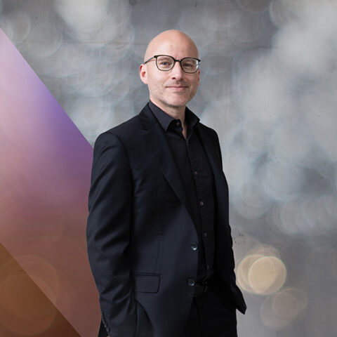 One man in a black suit with hands in pocket standing against grey background 
