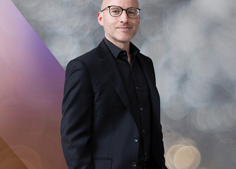One man in a black suit with hands in pocket standing against grey background 