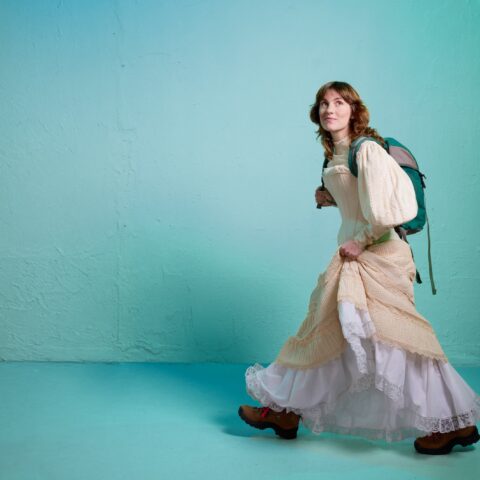 A young woman in a flowing cream dress walks across a turquoise wall with a backpack; soft lighting creates a dreamy, calm mood