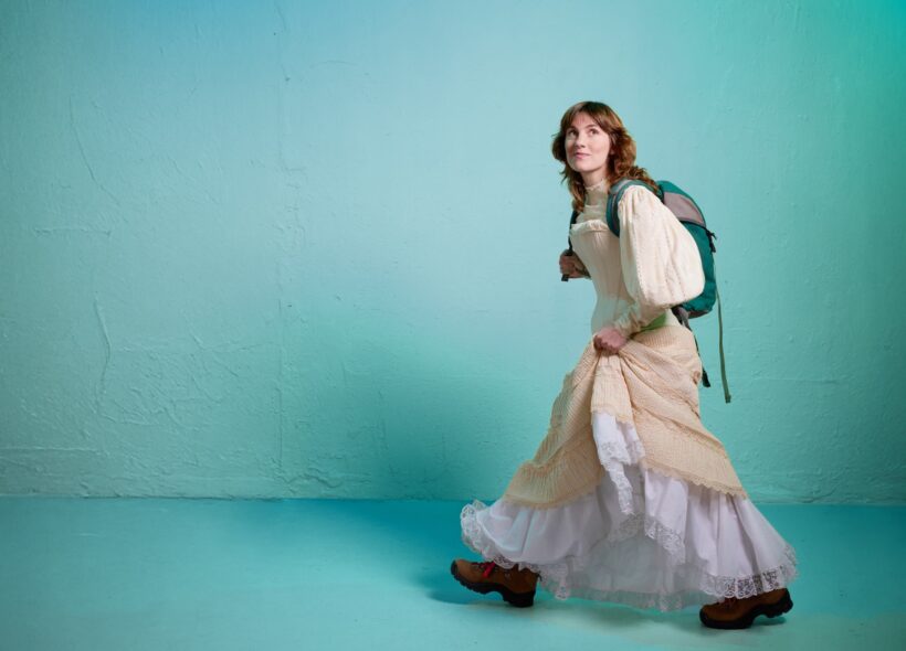 A young woman in a flowing cream dress walks across a turquoise wall with a backpack; soft lighting creates a dreamy, calm mood