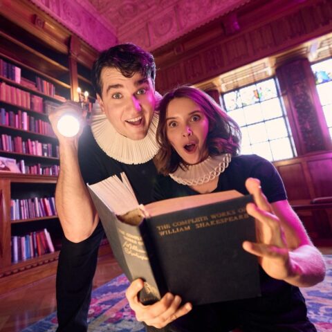 A man holding a flashlight and a woman with a surprised expression read from a large book in a library. They are both wearing ruffs around their necks