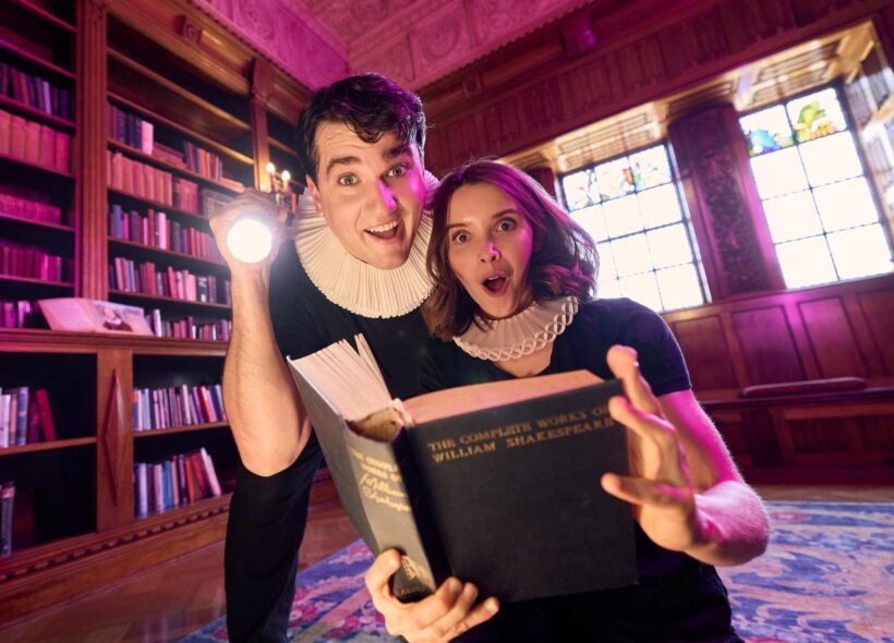 A man holding a flashlight and a woman with a surprised expression read from a large book in a library. They are both wearing ruffs around their necks