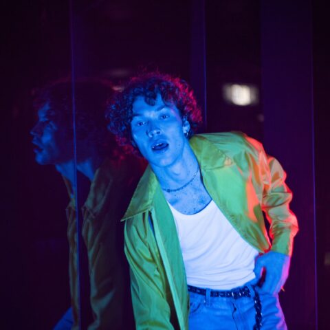Man wearing white singlet, green jacket and jeans poses with blue light washing over him.  