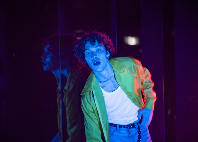 Man wearing white singlet, green jacket and jeans poses with blue light washing over him.  