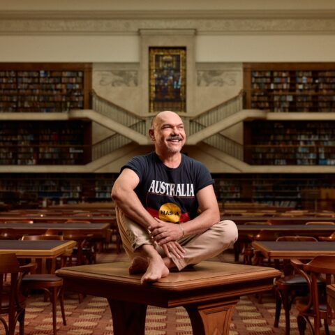 A man seated with legs crossed a wooden table in a library, surrounded by books and shelves wearing an Aboriginal flag on his shirt