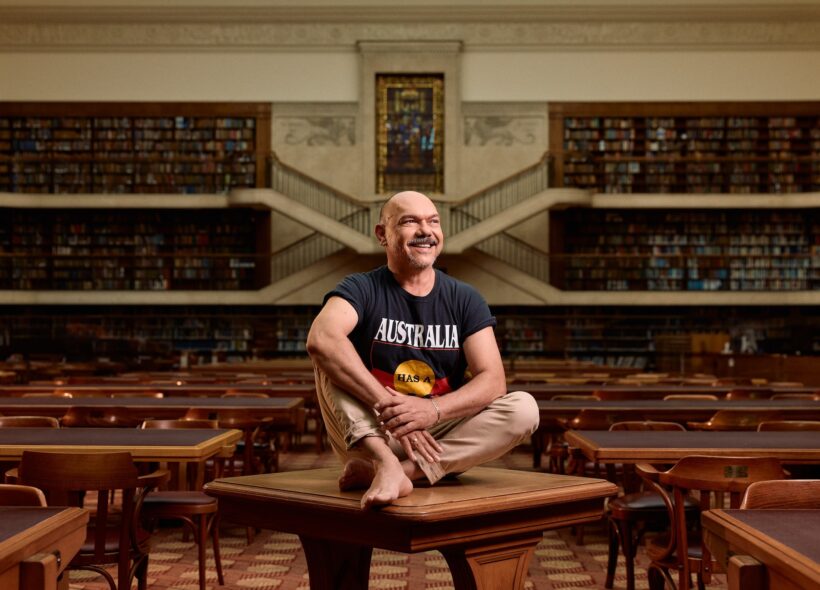 A man seated with legs crossed a wooden table in a library, surrounded by books and shelves wearing an Aboriginal flag on his shirt
