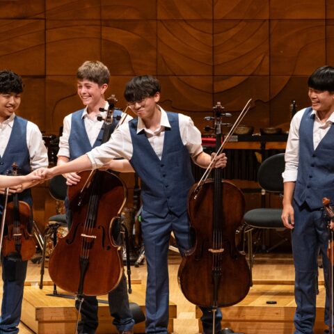A group of young musicians joyfully playing violins and a cello together in a bright, lively setting